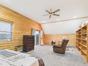 Une chambre de style rustique, aménagée dans une cabane en rondins, comprenant un lit, une chaise, une commode, un ventilateur de plafond et une fenêtre avec des stores.