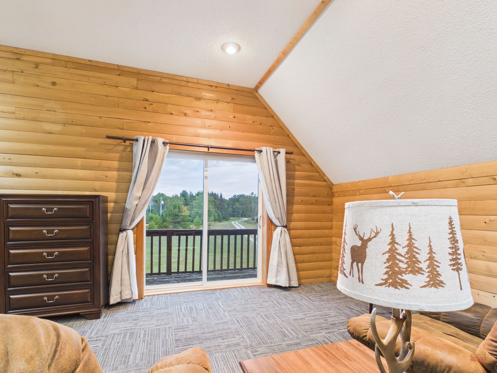 A cozy room with wooden walls, a large window with curtains, a wooden dresser, a sofa, and a lamp with a forest design.