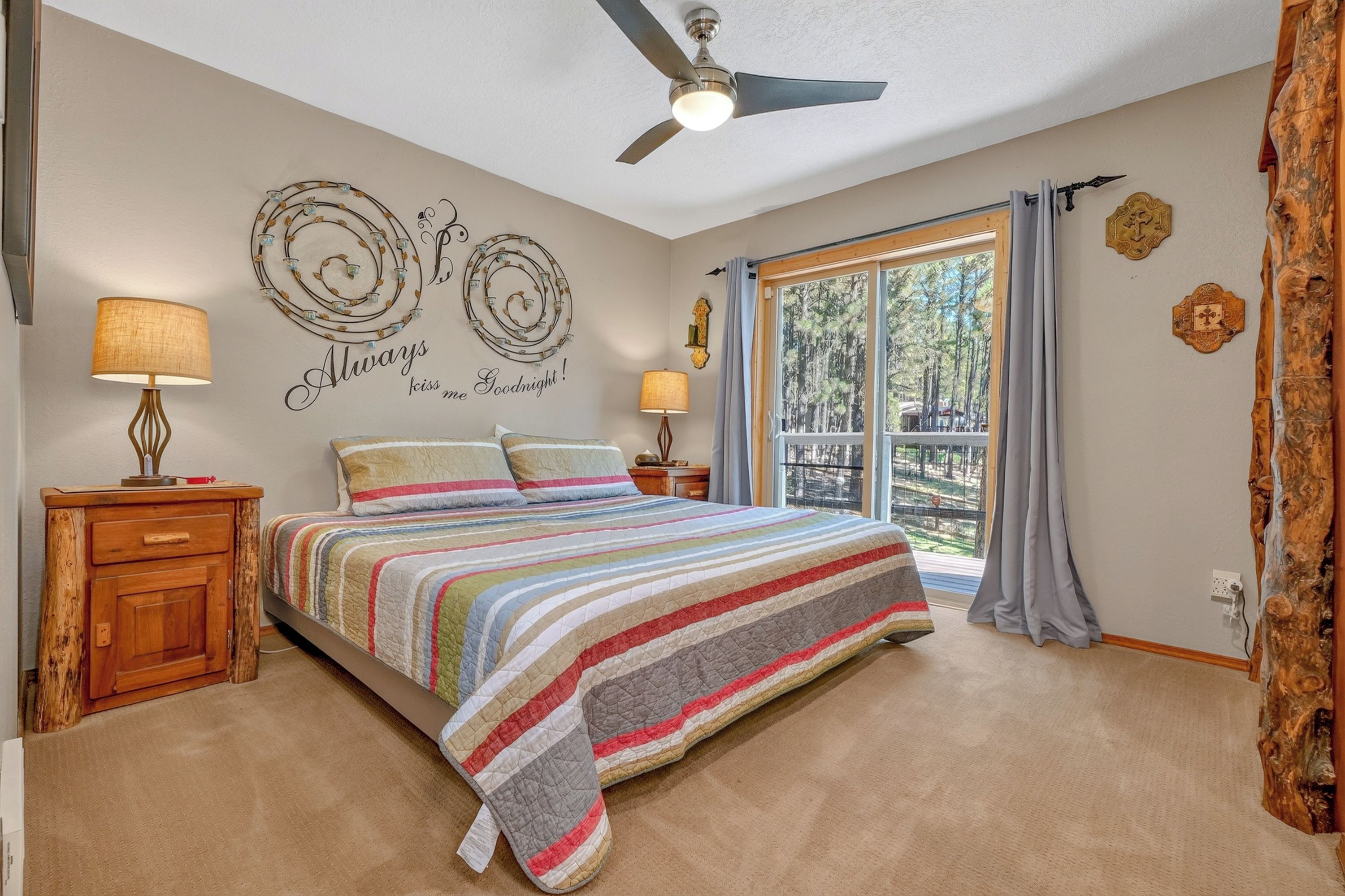A bedroom with a bed, two bedside lamps, a ceiling fan, a window with curtains, and a wall with decorative text.