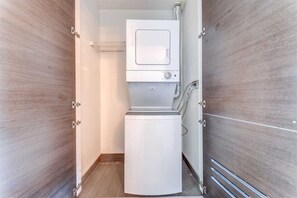 Une buanderie moderne et compacte, dotée d’un lave-linge et d’un sèche-linge intégrés, entourée de murs en panneaux de bois et équipée d’une étagère métallique au-dessus du sèche-linge.