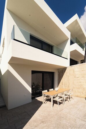 A modern two-story building with a flat roof, a balcony, and a patio area with a wooden table and chairs. - A Cube Villa - Two Bedroom Villa, Sleeps 5 (Keri)