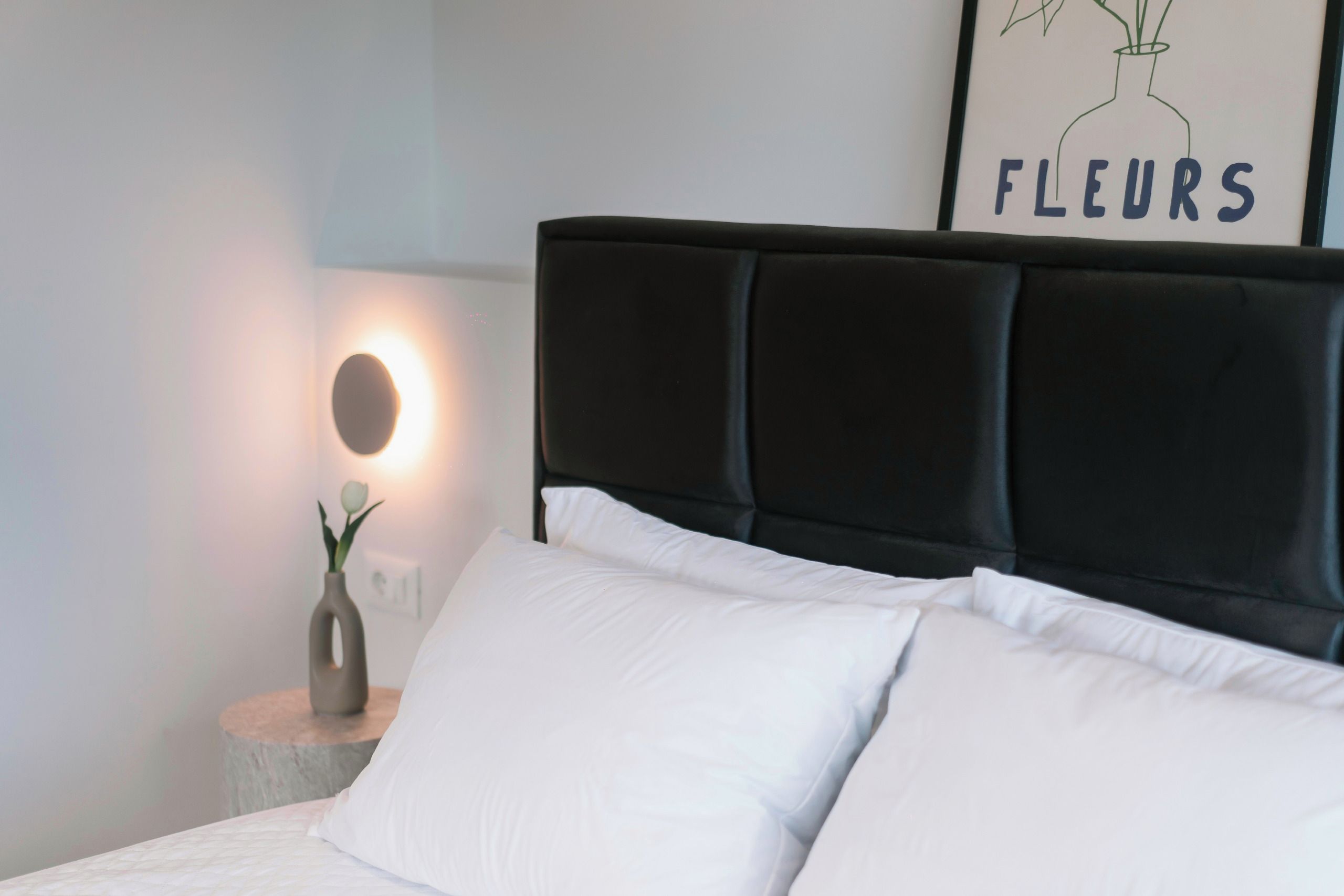 A bed with a black headboard and white pillows, a bedside table with a vase of flowers, and a wall-mounted light.