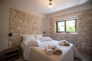 A bedroom with a stone wall, a bed with white bedding, a bedside table, and a window with a view of greenery. - Avesta Private Villas Kallisto (Lefkada)