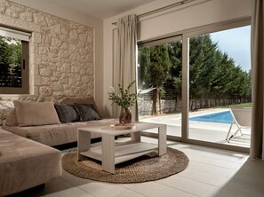 Ein modernes Wohnzimmer mit Steinwand, einem beigen Sofa, einem Couchtisch und Blick auf einen Swimmingpool und Bäume.