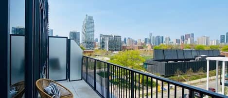 A balcony with a view of a city skyline, wicker chairs, a table, and a metal railing.