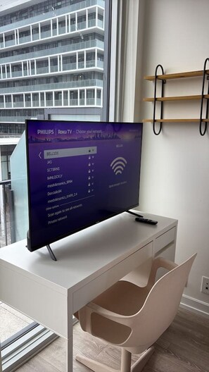 Une chambre moderne avec un téléviseur à écran plat posé sur un bureau blanc, une chaise et une fenêtre donnant sur un immeuble.