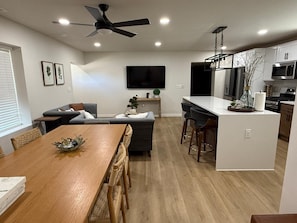 A modern kitchen with a central island, a dining area with a wooden table and chairs, a ceiling fan, recessed lighting, and a flat-screen TV mounted on the wall.
