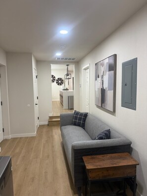 A modern living room with a grey sofa, a wooden coffee table, and a hallway leading to a kitchen area.