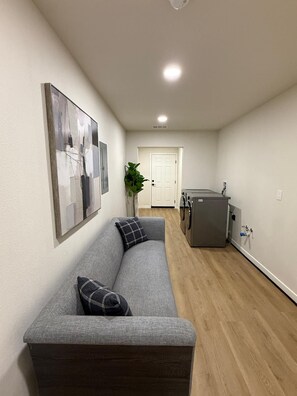 A modern living room with a gray sofa, a large abstract painting, a potted plant, and a kitchen area with a gray refrigerator. - Modern 3-Bedroom Home with Garage, Smart TVs, and Laundry Facilities. (Merced)