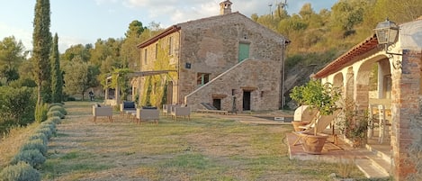A stone building with a staircase leading to a green door, surrounded by outdoor seating and a garden.