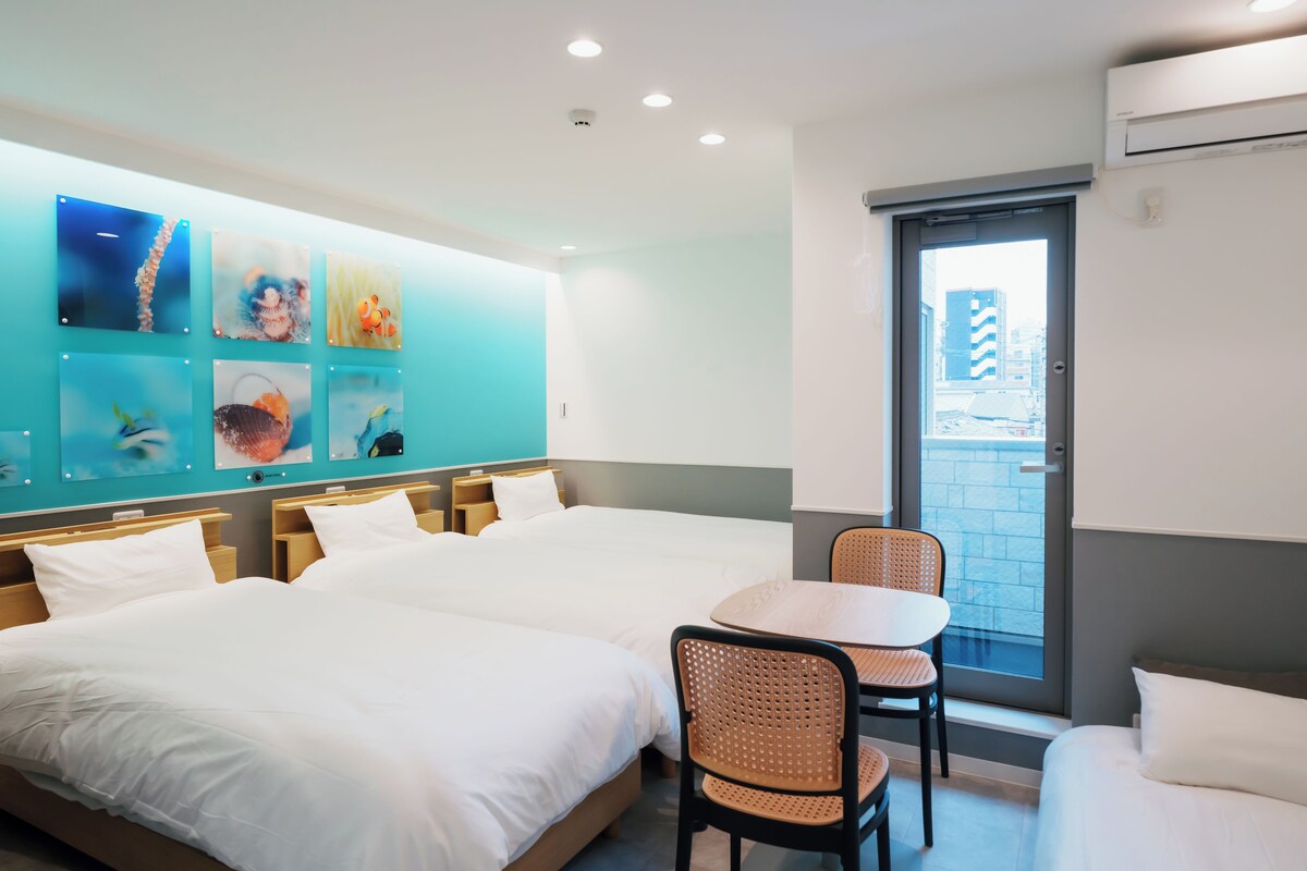 A hotel room with two beds, a small table, and chairs. There are wall-mounted artworks and a window with a view of buildings.