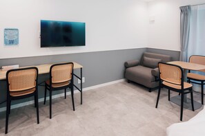 A hotel room with a flat-screen TV mounted on the wall, a desk with chairs, a sofa, and a window with curtains. - Ocean Blue 9 (Osaka)