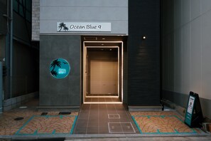 The entrance to Ocean Blue 9 with a closed shutter door, a sign with palm tree and bird silhouettes, and a tiled pavement with directional markings. - Ocean Blue 9 (Osaka)