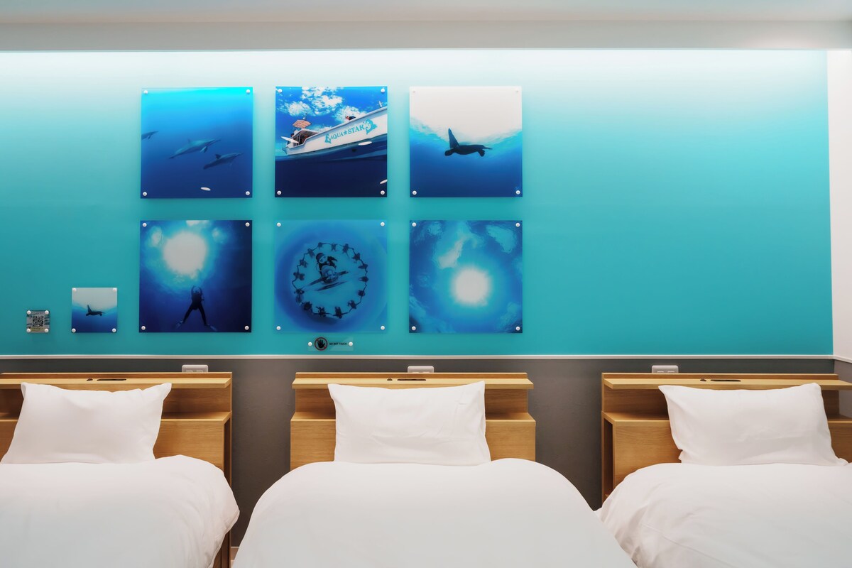 A hotel room with a wall of six framed aquatic-themed artworks, two beds, and a bedside table with a phone.
