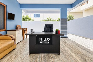 A hotel reception desk with a sign indicating OYO-Serviced, a wooden sofa, a chair, and a small table with a red cup.