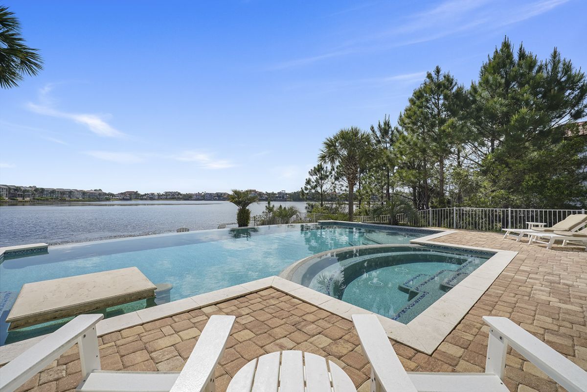 Une piscine donnant sur un plan d’eau, entourée d’une terrasse en briques et de chaises longues blanches.