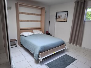 A bedroom with a bed, a bedside table, a nightstand, a wall-mounted lamp, a framed picture, and a window with curtains. - Vrbo Property (Seclin)