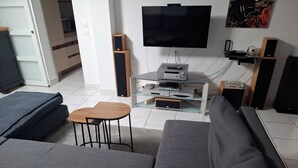 A modern living room with a grey sofa, a small wooden table, a flat-screen TV, a sound system, and a glass entertainment unit. - Vrbo Property (Seclin)
