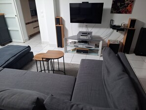 Uma sala de estar moderna com um sofá cinzento, uma pequena mesa de madeira, uma TV de tela plana, um sistema de som e um rack para eletrônicos de vidro.