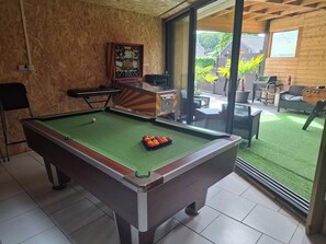 Uma mesa de bilhar com bola branca e bolas vermelhas e amarelas, um quadro na parede e vista para um pátio ao ar livre com móveis e churrasqueira.