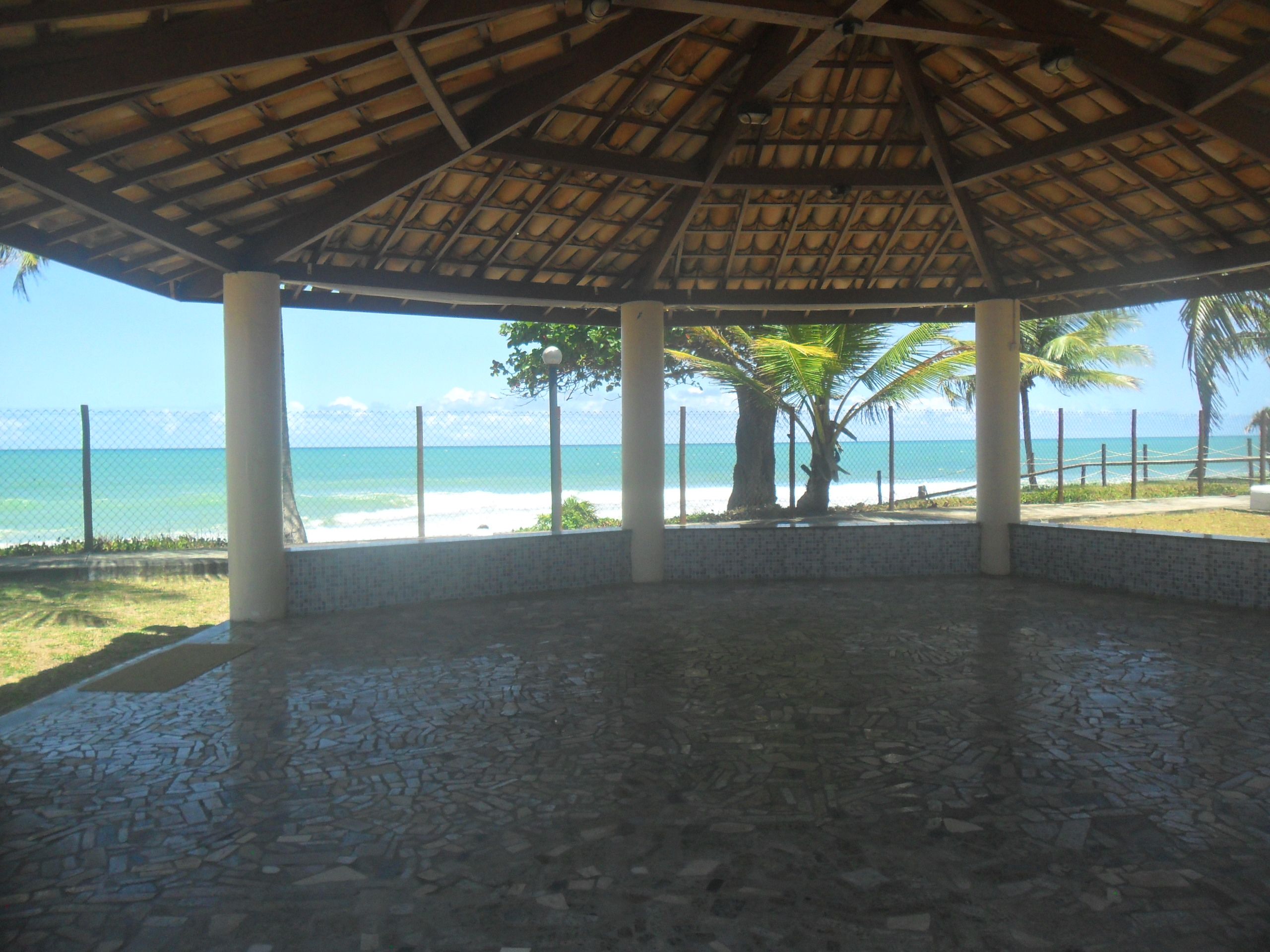 Uma área externa coberta com piso de azulejos, palmeiras e vista para a praia e o oceano.