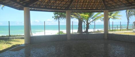 Un espacio al aire libre techado con piso de baldosas, palmeras y vista a la playa y al océano.