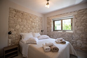 Ein Schlafzimmer mit Steinwand, einem Bett mit weißer Bettwäsche, einem Nachttisch und einem Fenster mit Blick auf Grünflächen.