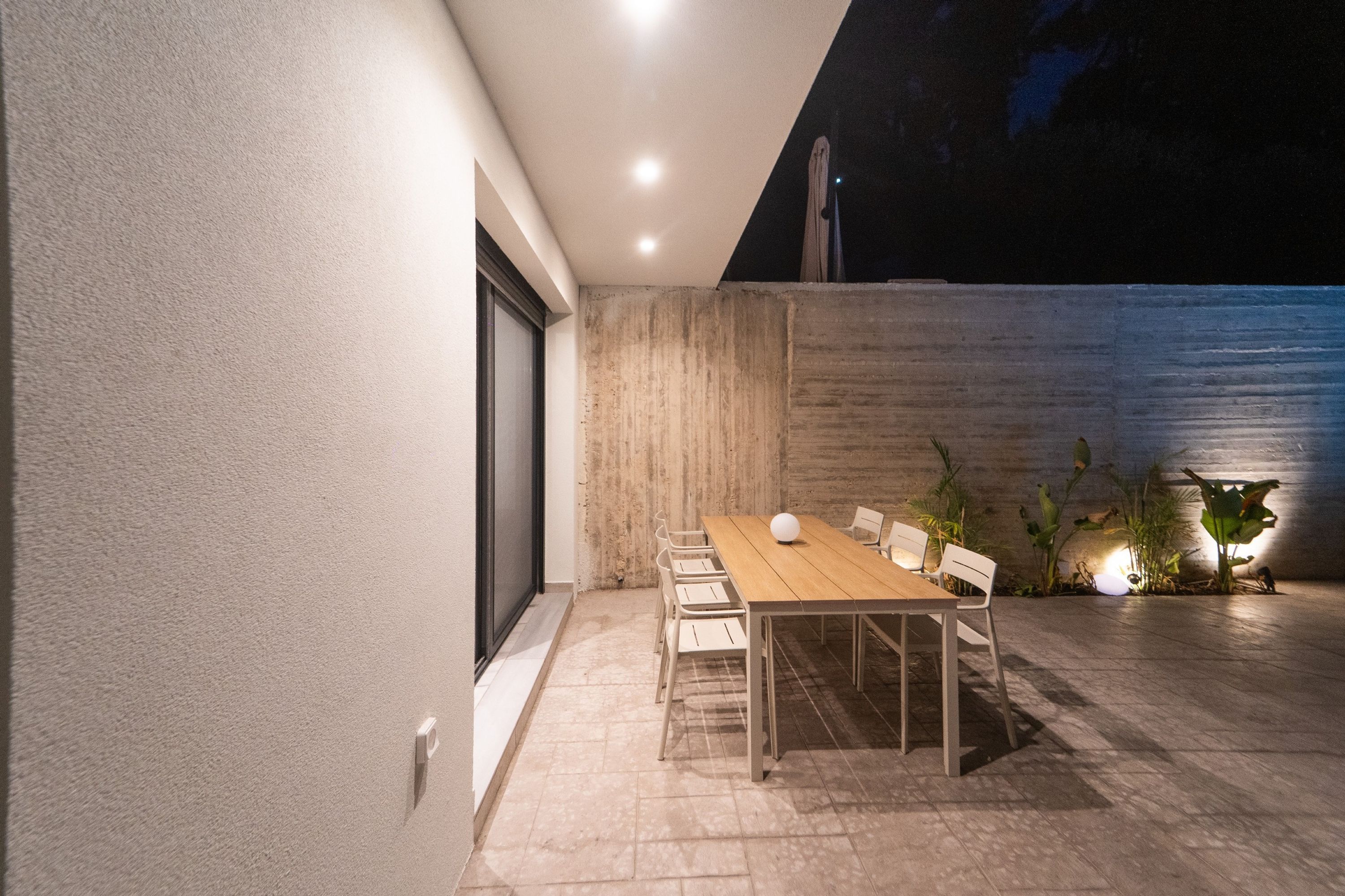 An outdoor patio with a wooden table and chairs, a concrete wall, and a small garden area with plants.