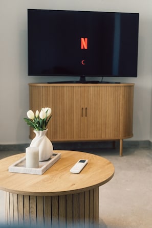 Ein Zimmer mit einem Holzschrank, einem Fernseher mit Netflix-Logo, einer weißen Vase mit Blumen auf einem runden Holztisch und einem Smartphone auf dem Tisch.