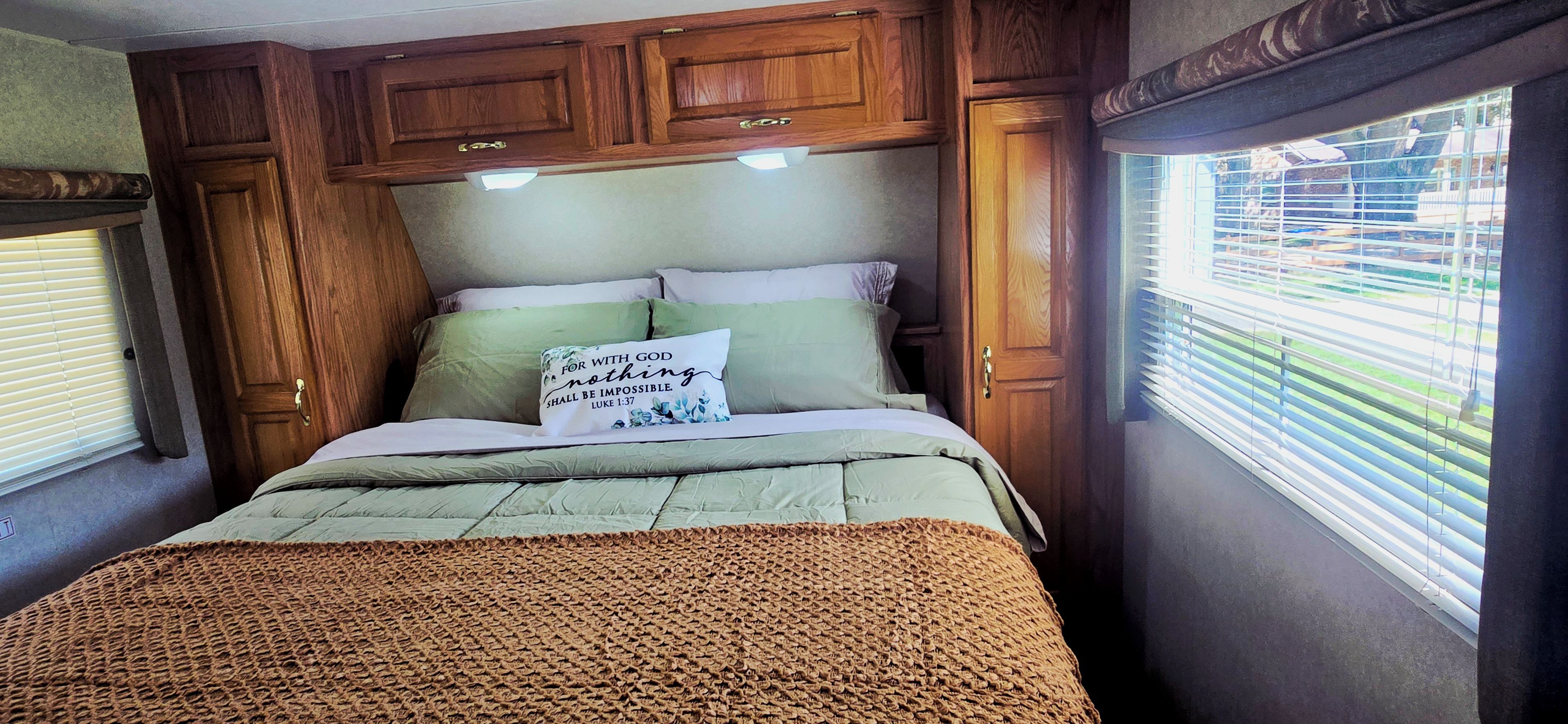 A bedroom with a bed, pillows, a blanket, wooden cabinets, and a window with blinds.