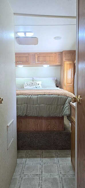 A compact RV interior with a bed, wooden cabinets, and a tiled floor. - Serene 1-bedroom, 1-bathroom, 5th wheel with AC, farms next-door near Lucedale. (Lucedale)