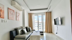 A modern living room with a sofa, a flat-screen TV mounted on the wall, and a large window with curtains. - EMERALD CELADON CITY 2PN2WC (Thanh pho Chi Minh)