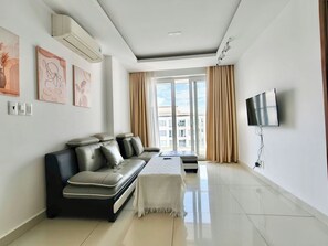 A modern living room with a sofa, a flat-screen TV mounted on the wall, and a large window with curtains. - EMERALD CELADON CITY 2PN2WC (Thanh pho Chi Minh)
