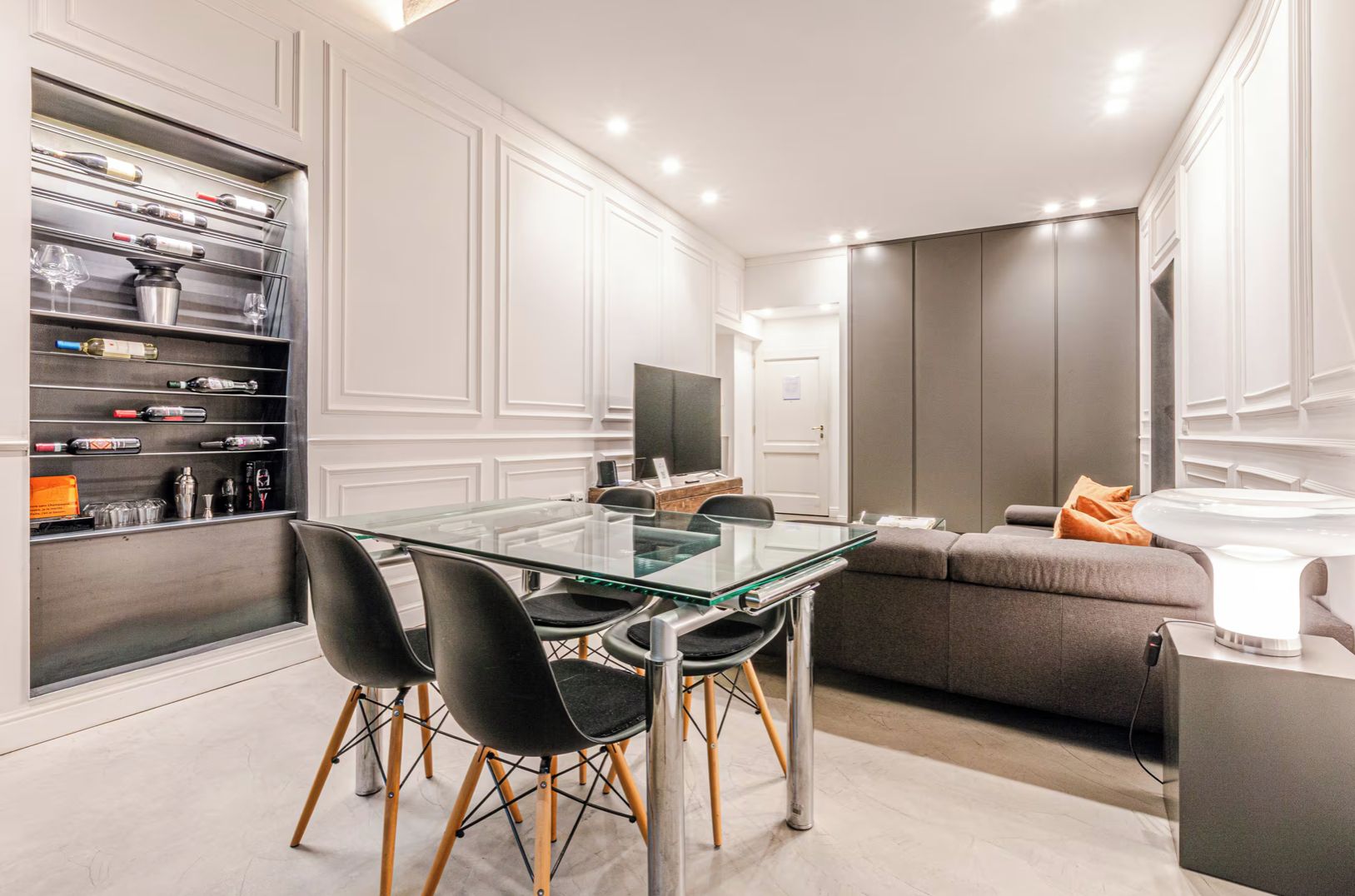 Un moderno comedor con mesa y sillas de vidrio, una estantería empotrada para vinos y un sofá con una mesita auxiliar.