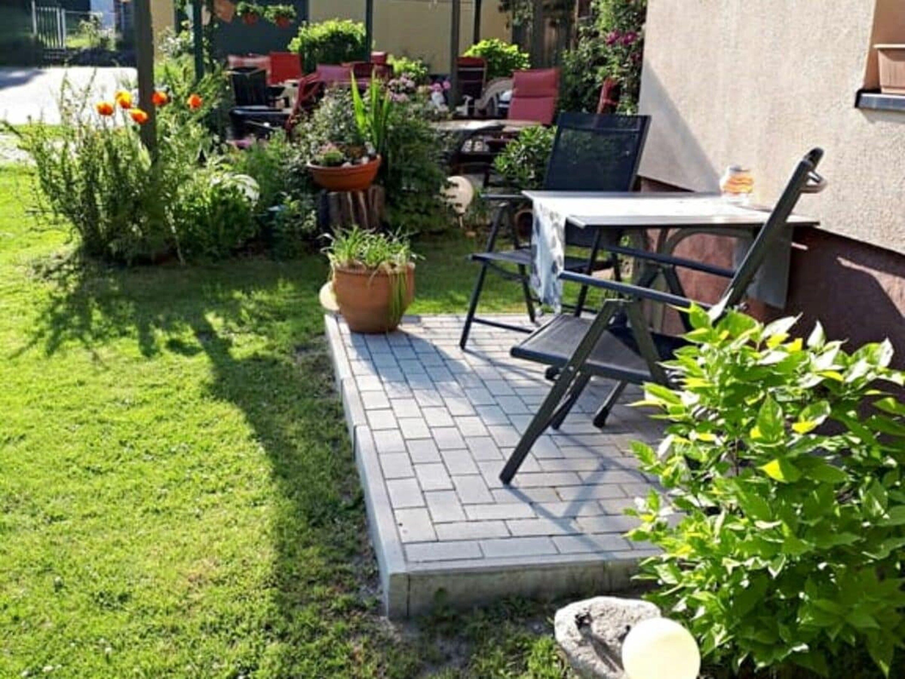An outdoor patio area with a table and chairs, surrounded by potted plants and a grassy lawn.