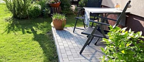 Un patio al aire libre con una mesa y sillas, rodeado de plantas en macetas y un césped.