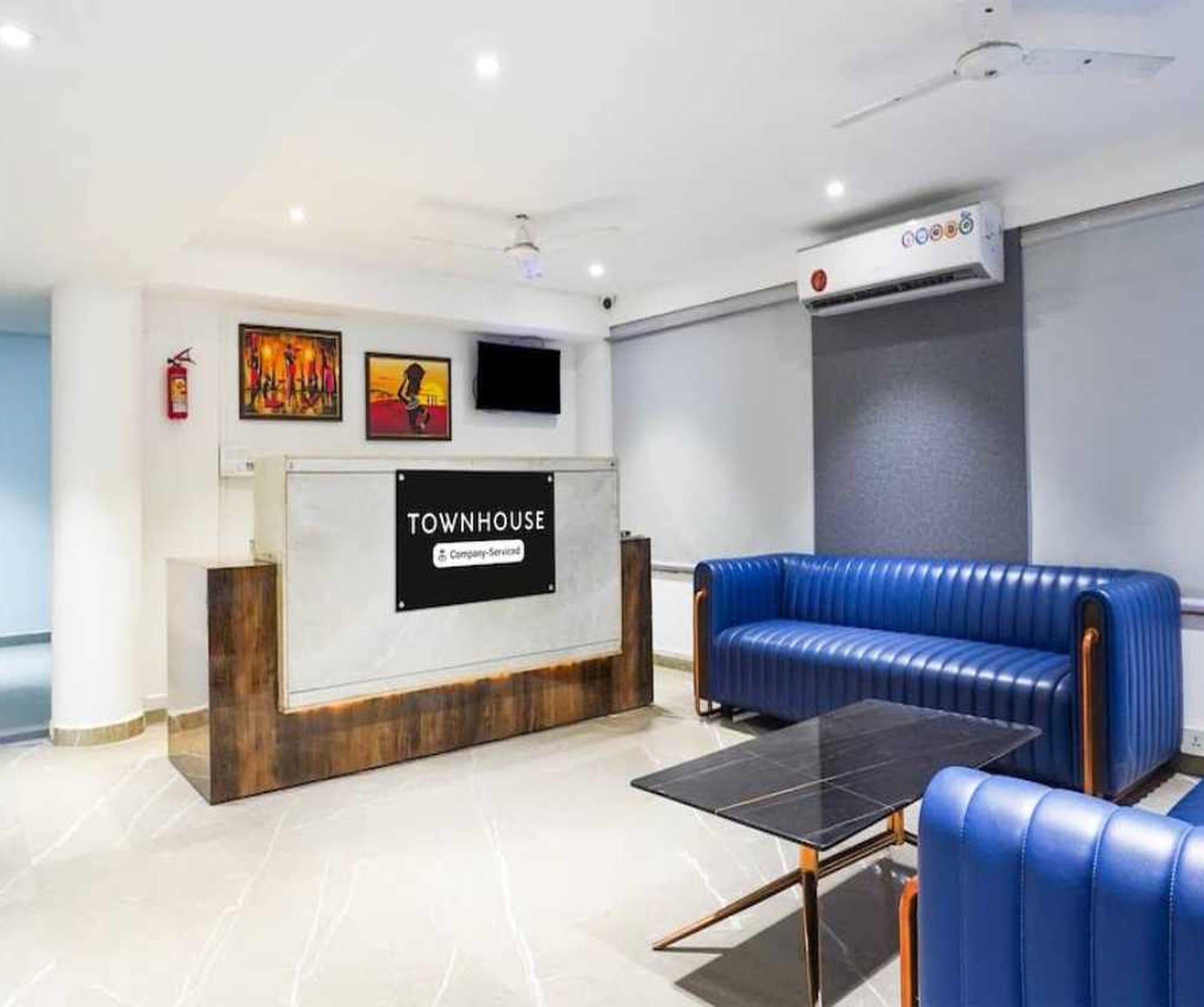 A reception area with a sign reading 'TOWNHOUSE,' a blue leather couch, a small table, and a mounted flat-screen TV.