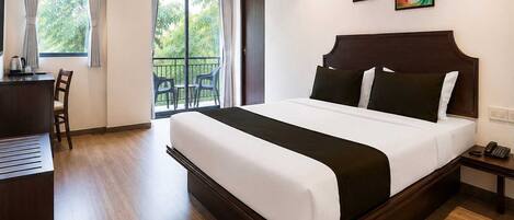 A hotel room with a large bed, a desk, a chair, a balcony with a view of greenery, and two paintings on the wall.