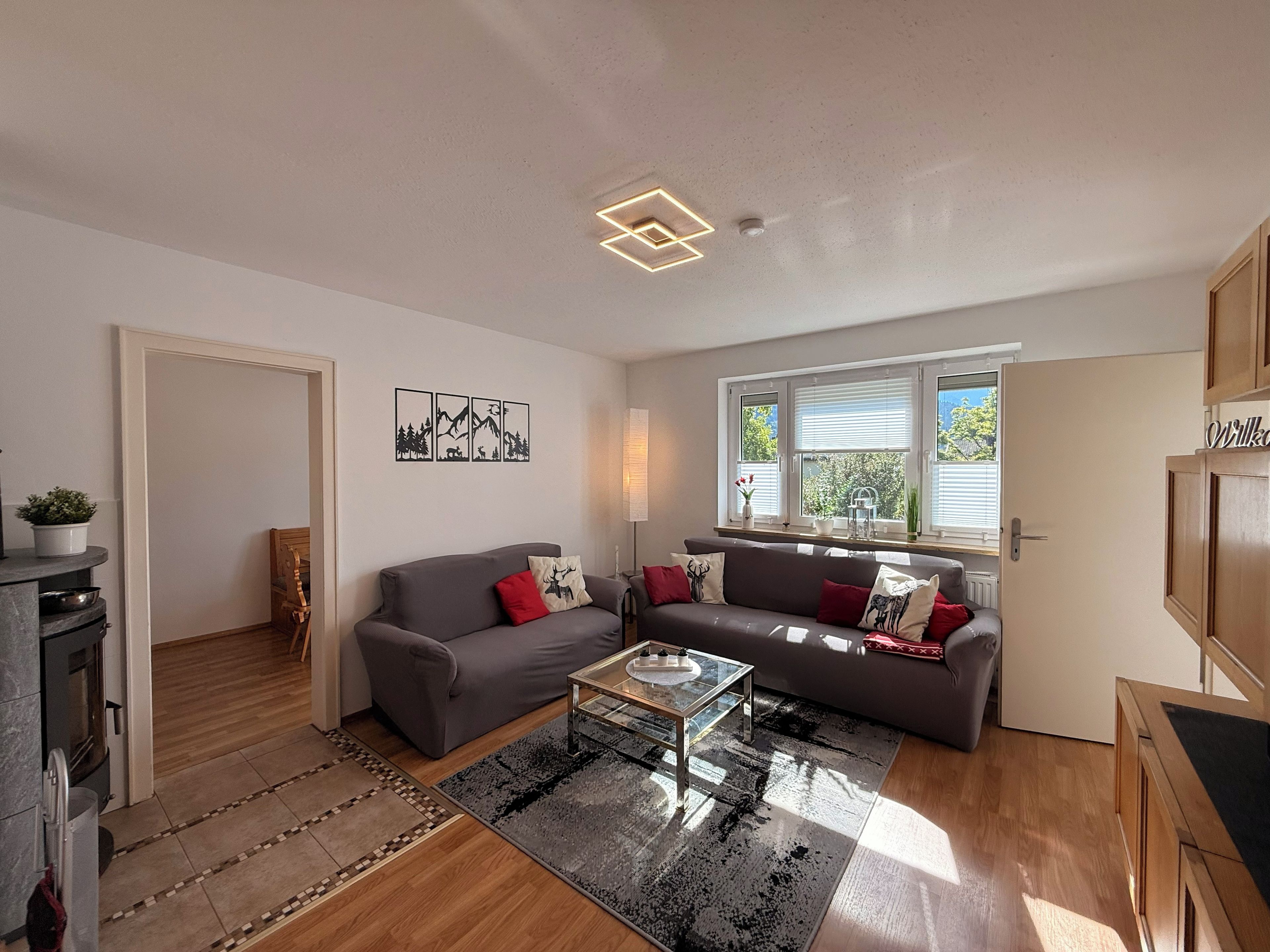 Ein modernes Wohnzimmer mit einem grauen Sofa, einem Couchtisch und einem Kamin. An der Wand hängen gerahmte Bilder und ein Fenster mit Jalousien.