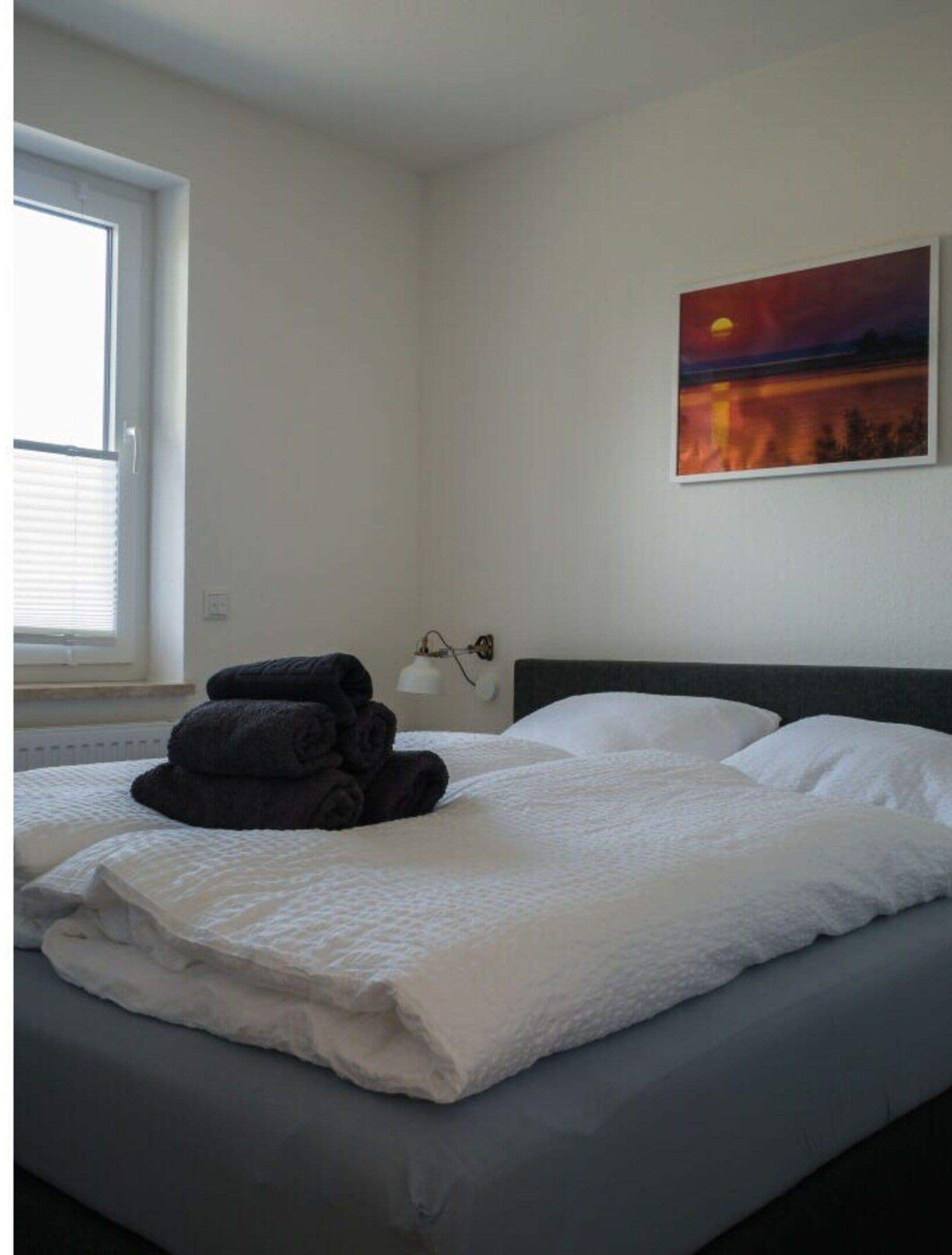 Una cama individual con ropa de cama blanca, cabecera oscura y un cuadro de un atardecer en la pared.
