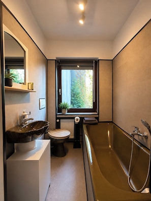 A modern bathroom with a freestanding bathtub, a sink, a toilet, and a window providing a view of greenery outside.