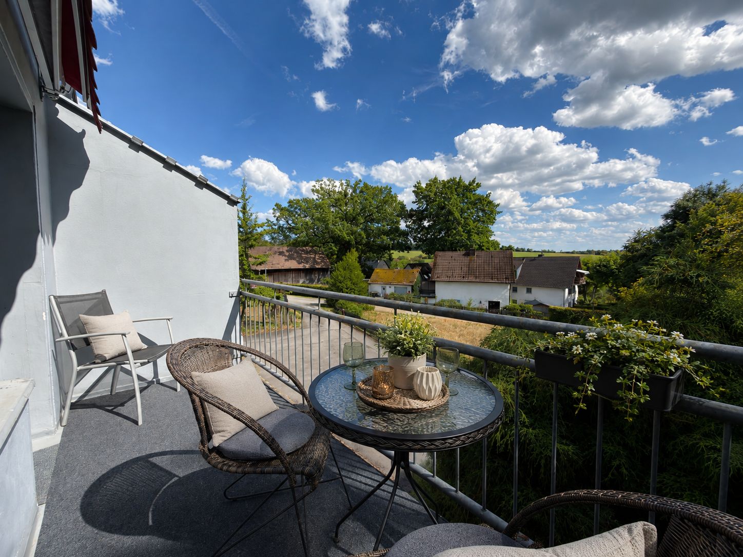 Ein Balkon mit Korbmöbeln, einem Glastisch und Blick auf Häuser und Grünflächen.