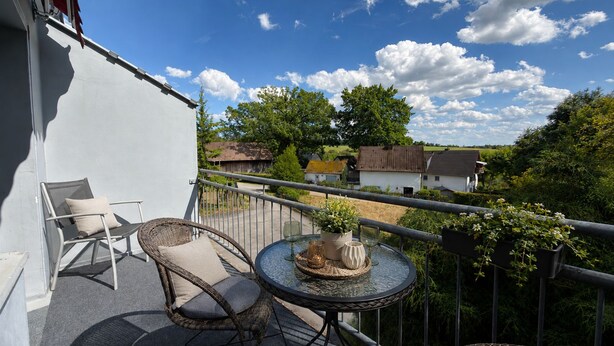 Varanda com móveis de vime, mesa de vidro e vista para casas e vegetação.