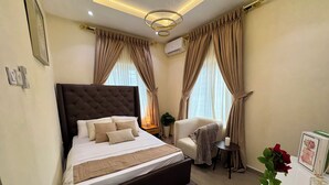 A hotel room with a large bed, a bedside table, a chair, a potted plant, and a desk with a vase of flowers. - Haven 21;  Luxury 1-Bedroom Serviced Apartment in Ajah (Close)