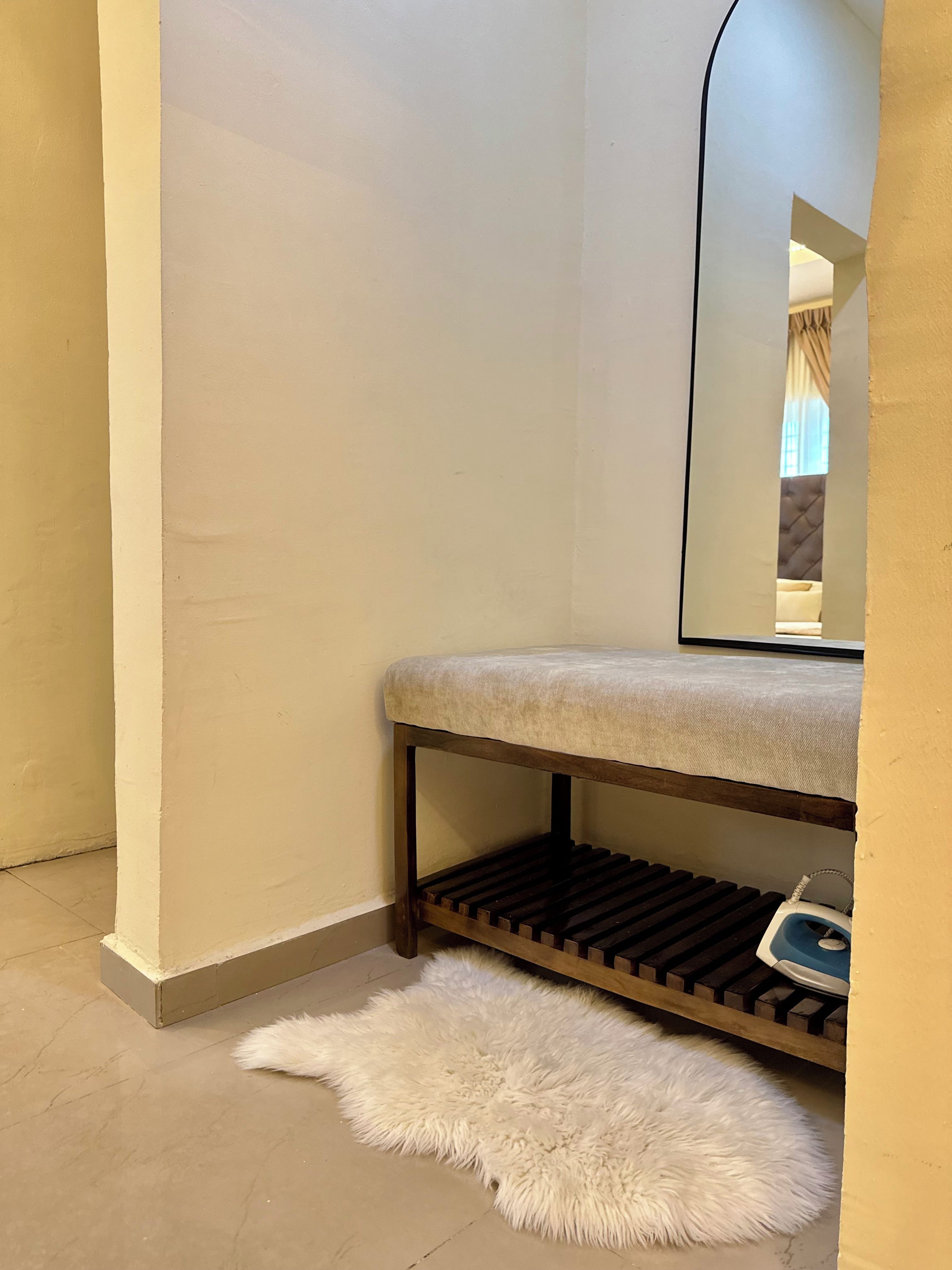 A corner of a room with a bench, a fluffy rug, and a mirror reflecting a window with curtains.