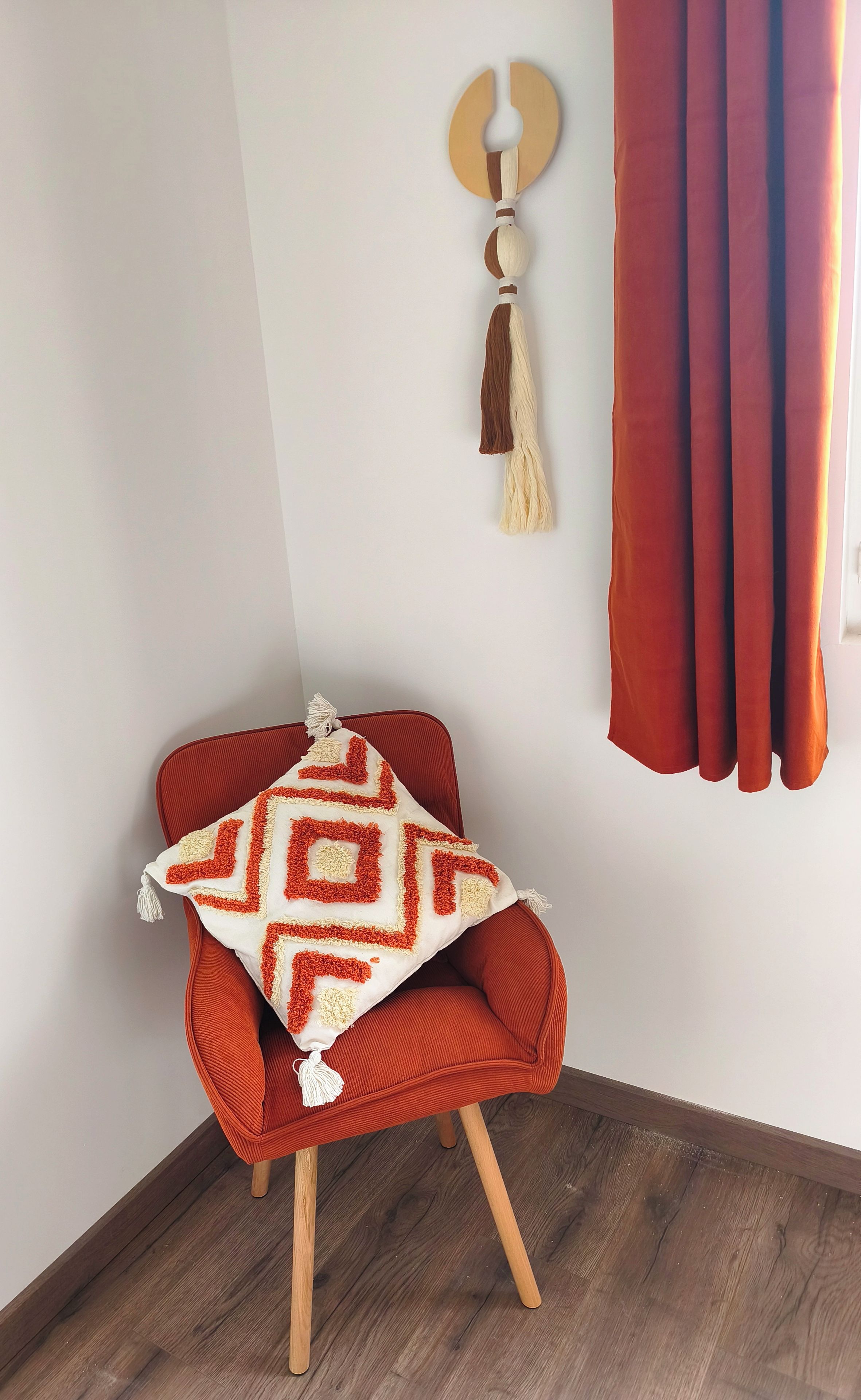 Une pièce d’angle avec un fauteuil rouge, un coussin décoratif à motifs, une frange décorative accrochée au mur et un rideau rouge.