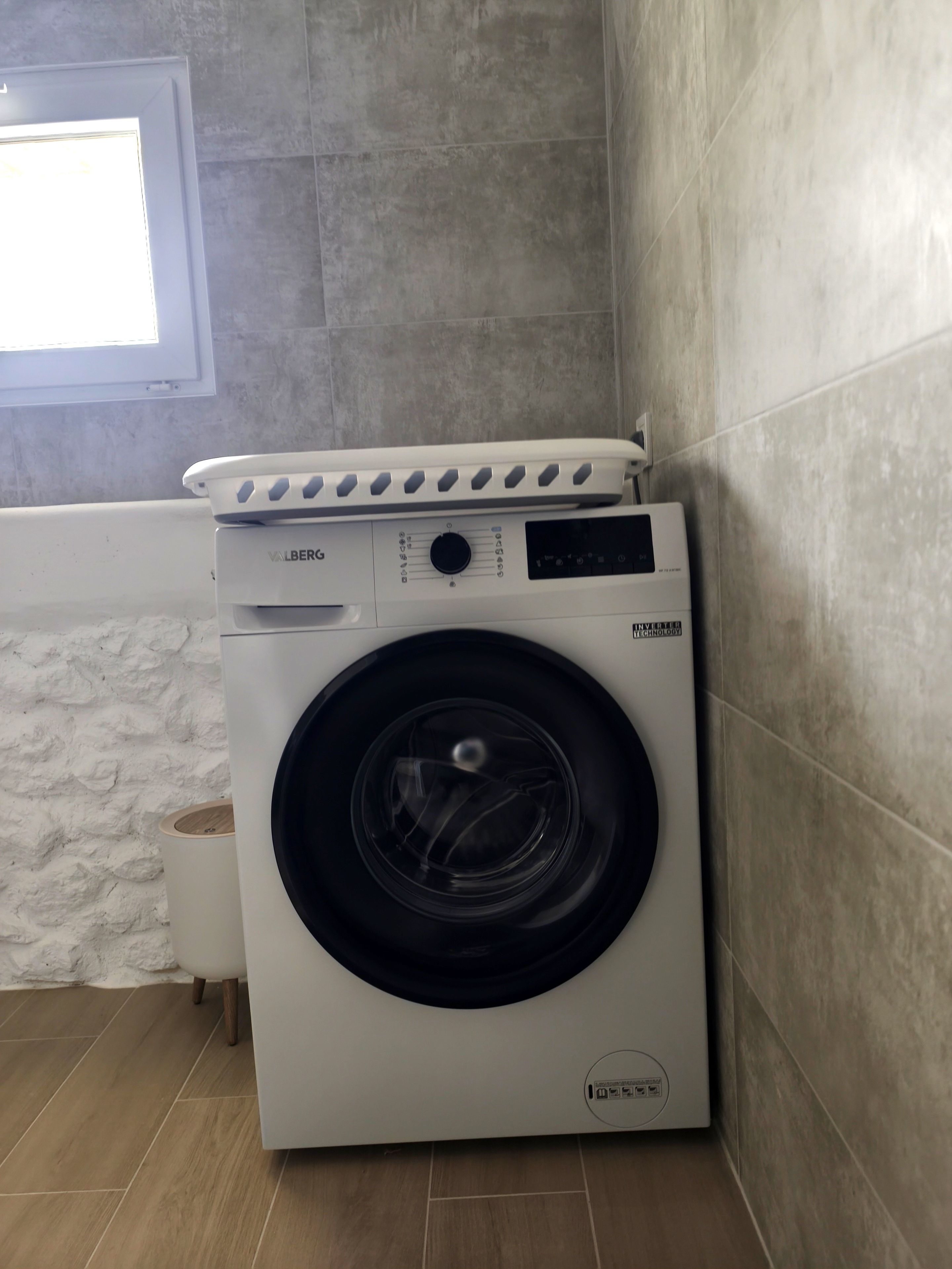Un lave-linge à chargement frontal doté d’un panneau de commande, situé dans un coin d’une pièce dont les murs sont recouverts de carreaux et qui possède une fenêtre.