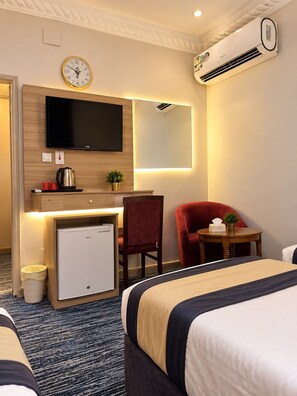 A hotel room with a bed, a desk with a television, a chair, a small refrigerator, a trash bin, and an air conditioning unit. - Sakab Al Aseel Hotel  (Makkah)