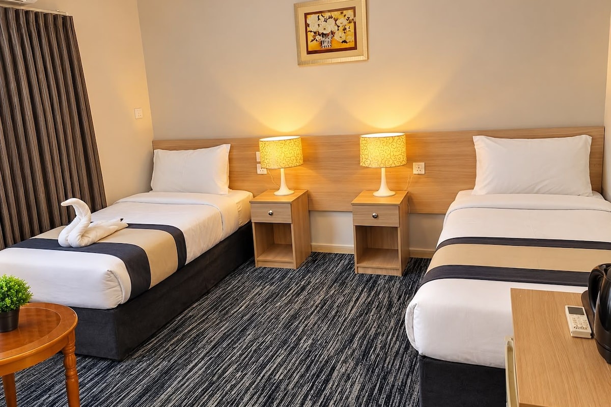 A hotel room with two beds, a small table, a chair, a potted plant, and a painting on the wall.