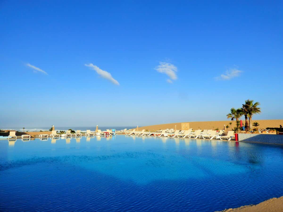 Une piscine d’un bleu éclatant, bordée de chaises longues et entourée de palmiers, avec une plage de sable fin et un ciel dégagé.